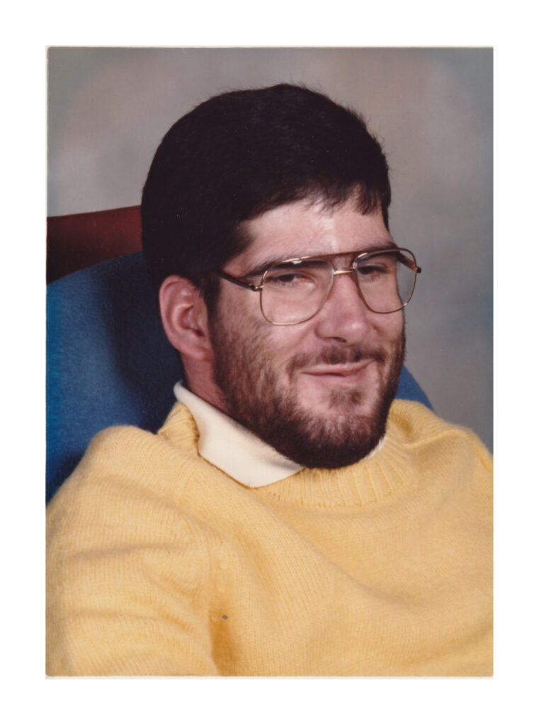 portrait of young man with dark hair and a beard wearing a yellow sweater.